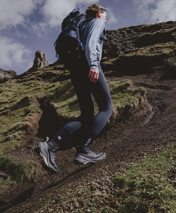 Wandernde Person mit Rucksack geht einen grasbewachsenen Pfad bergauf, dahinter Felsen und Wolken